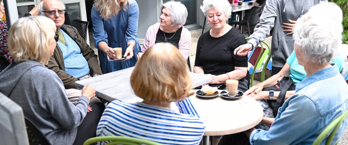 A group of older people sit around a table, they are being served coffee by two younger women. A sign with the words Ajani Community Hall can be seen on a window behind them.