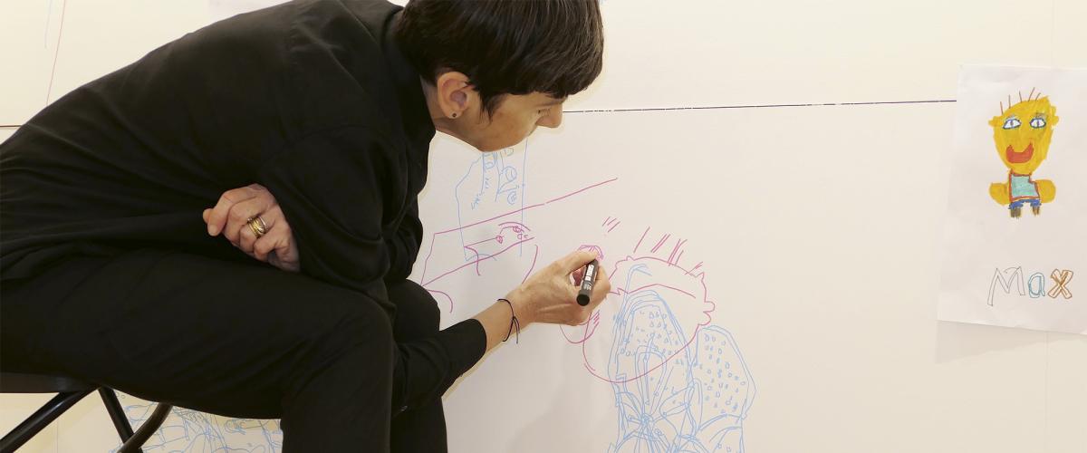 Woman dressed in black sits on stool and draws a face onto the wall, child's drawing with the word Max is stuck to the wall to her right