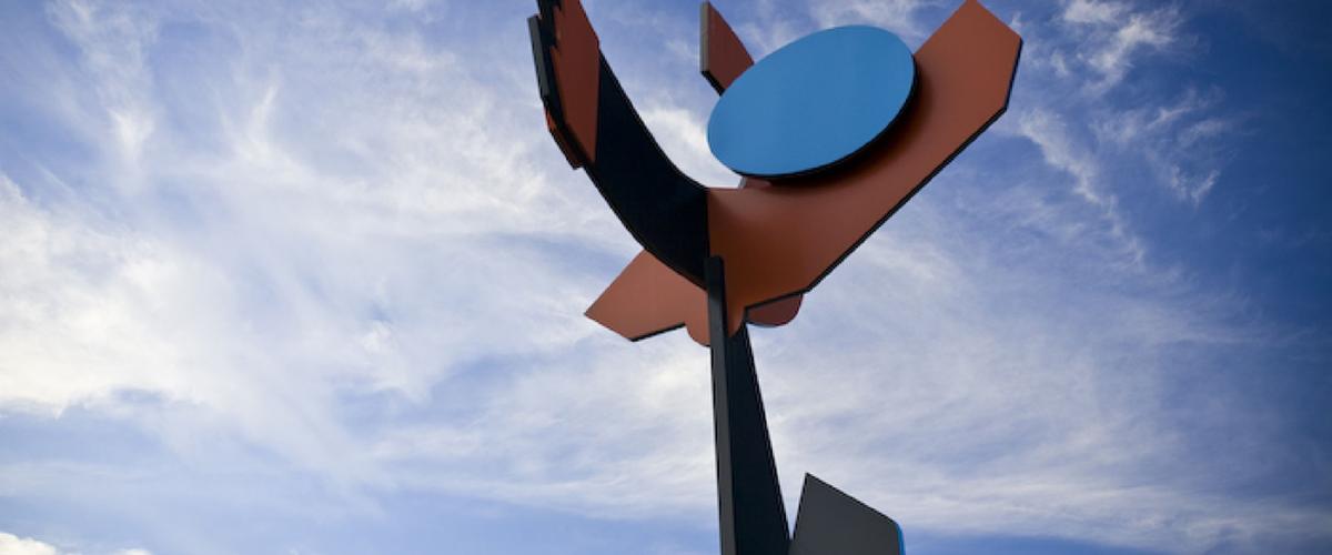 Large steel sculpture comprising bold geometric shapes in black, brown and blue sits against a blue sky.