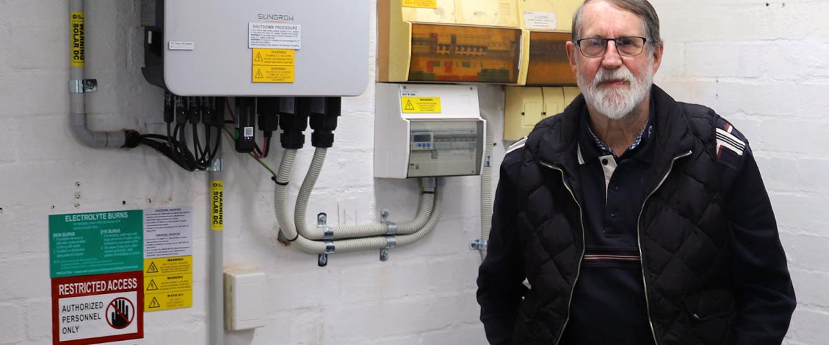 A man wearing glasses and a black jacket stands with his hands in his pockets. He is in a garage and a solar battery and a switchboard can be seen on the wall to his right.