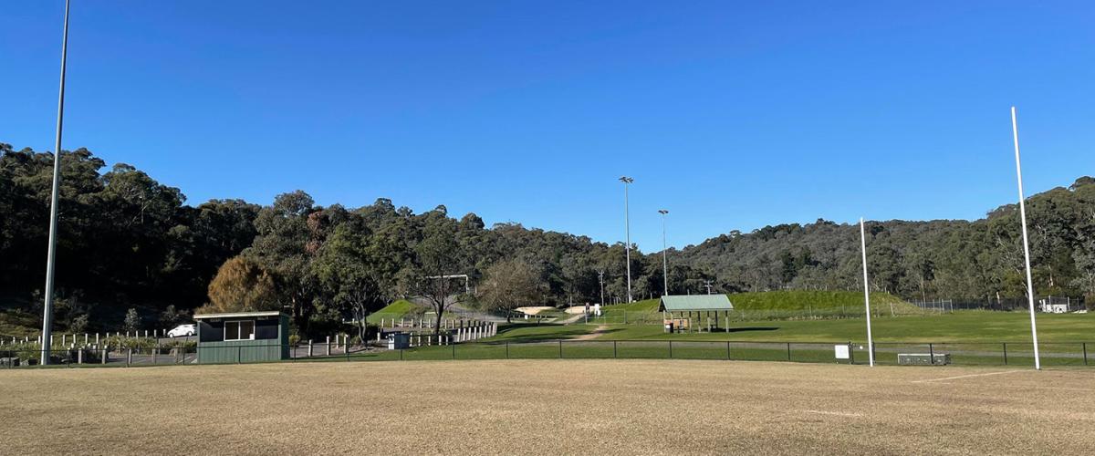 An empty football over with light poles and football goals around the outside, bushy parklands in the background.