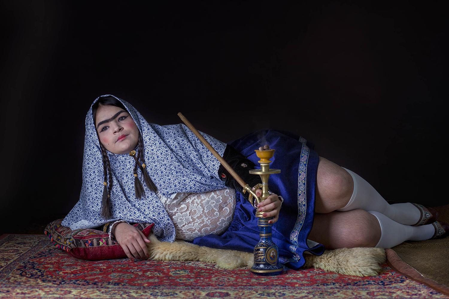 A young Iranian woman reclines on a red patterned carpet, she is holding a hookah pipe.