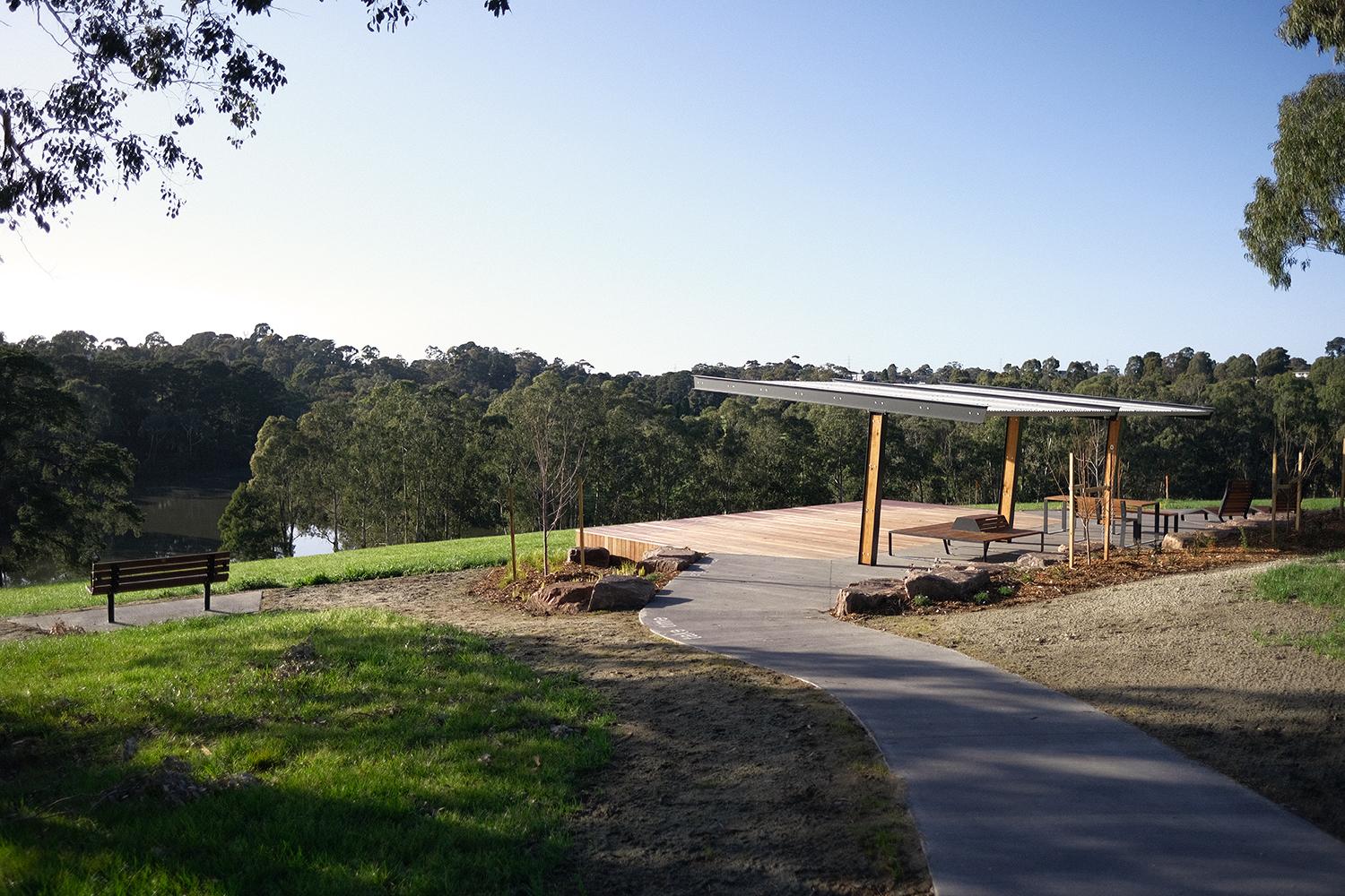 A concrete path leads to a wooden pavilion sitting at the top of a gently sloping hill looking towards bushland and a lake.