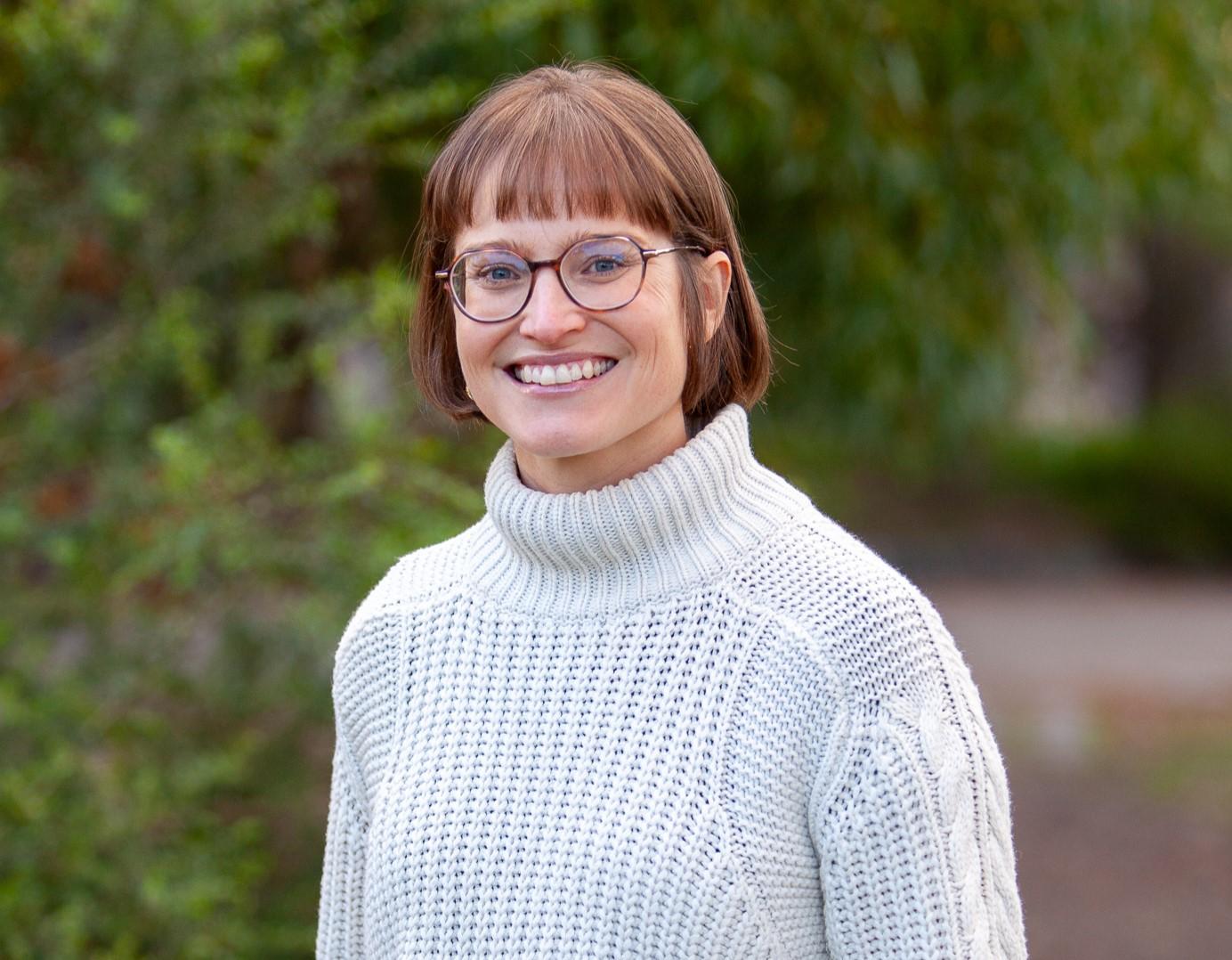 A woman stands smiling with her hands in her pockets, she has short brown hair and glasses