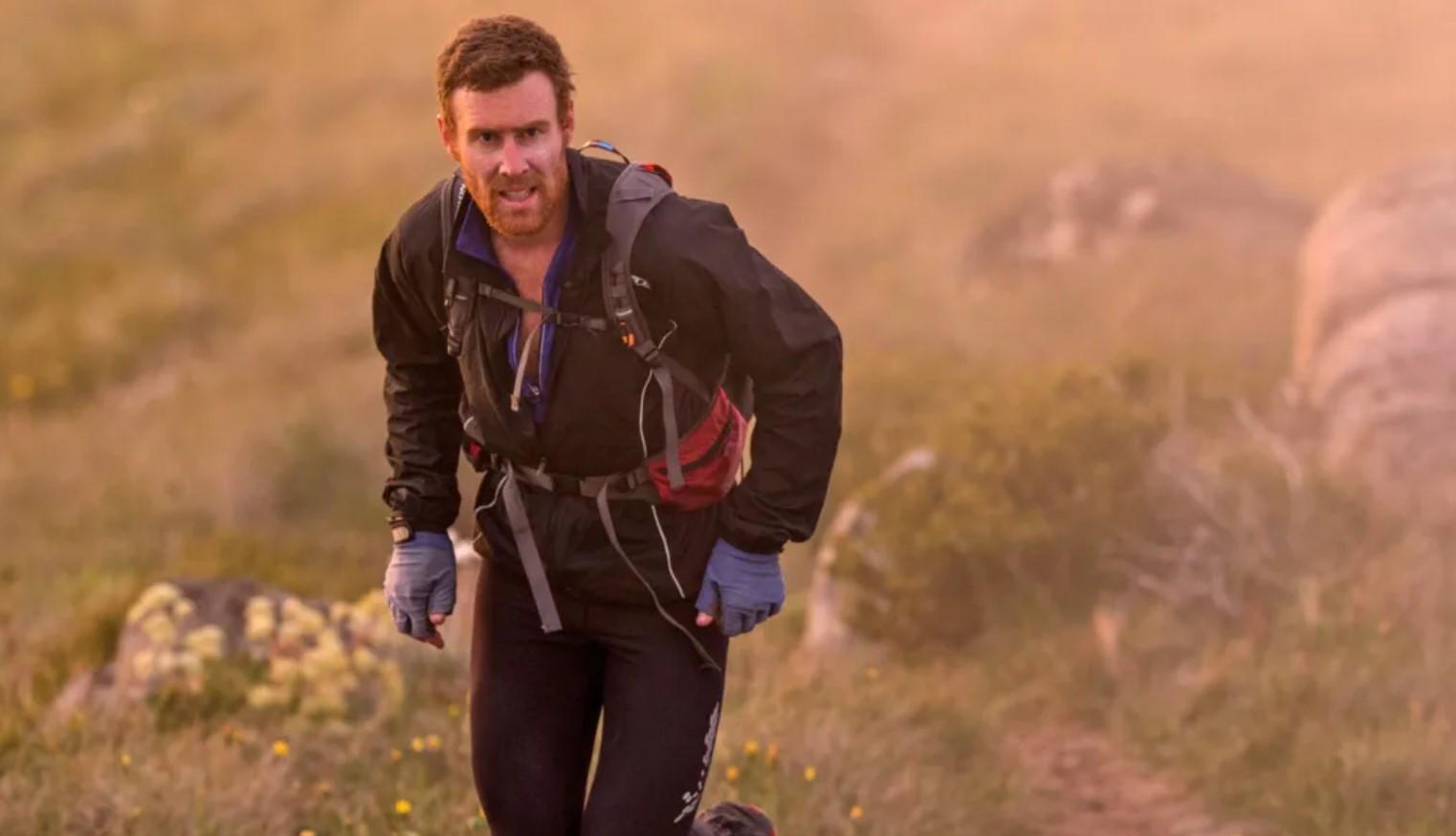 A man dressed in black and wearing a backpack and gloves runs up a mountain path