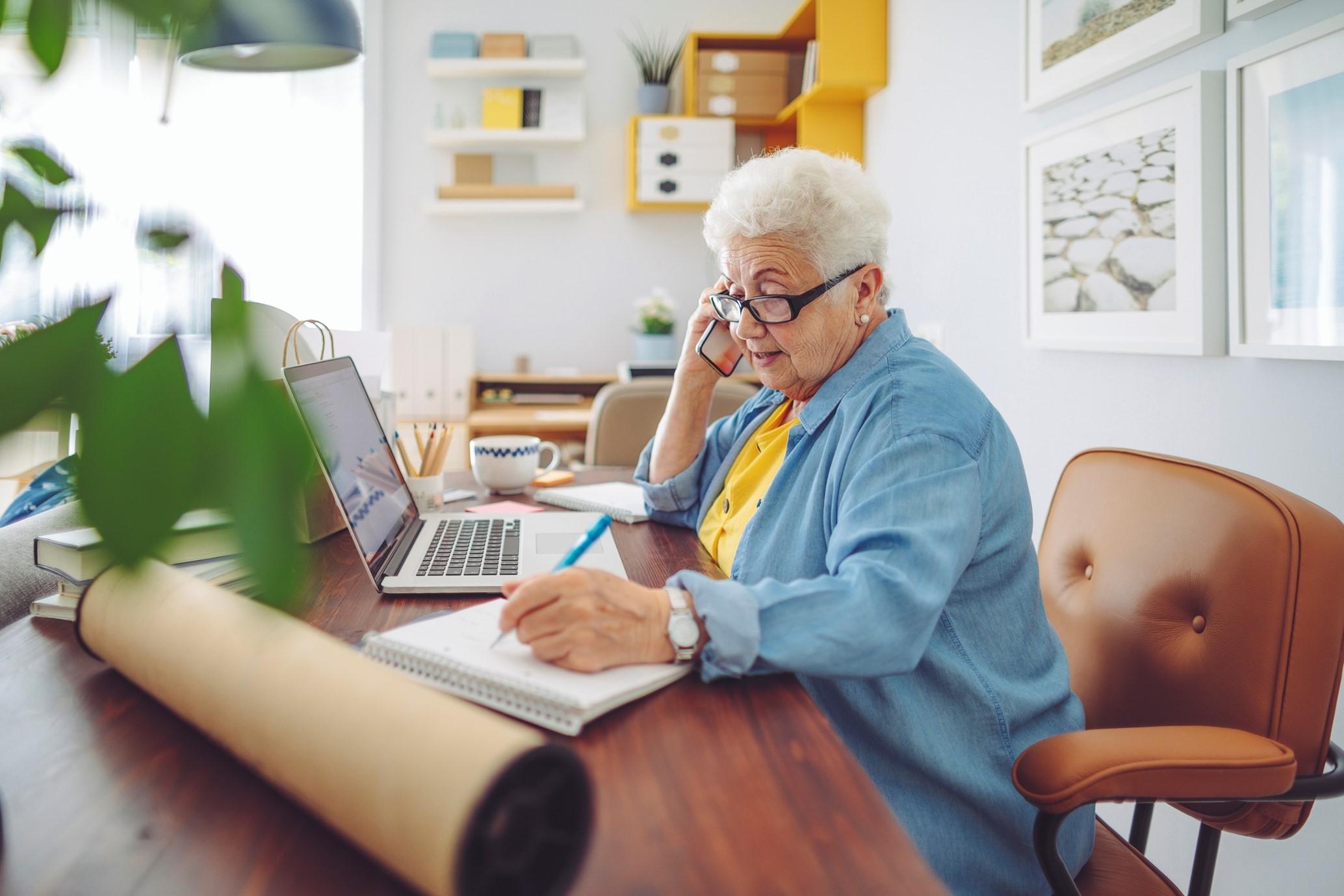 Older woman writing with pen while on the phone in front of a laptop