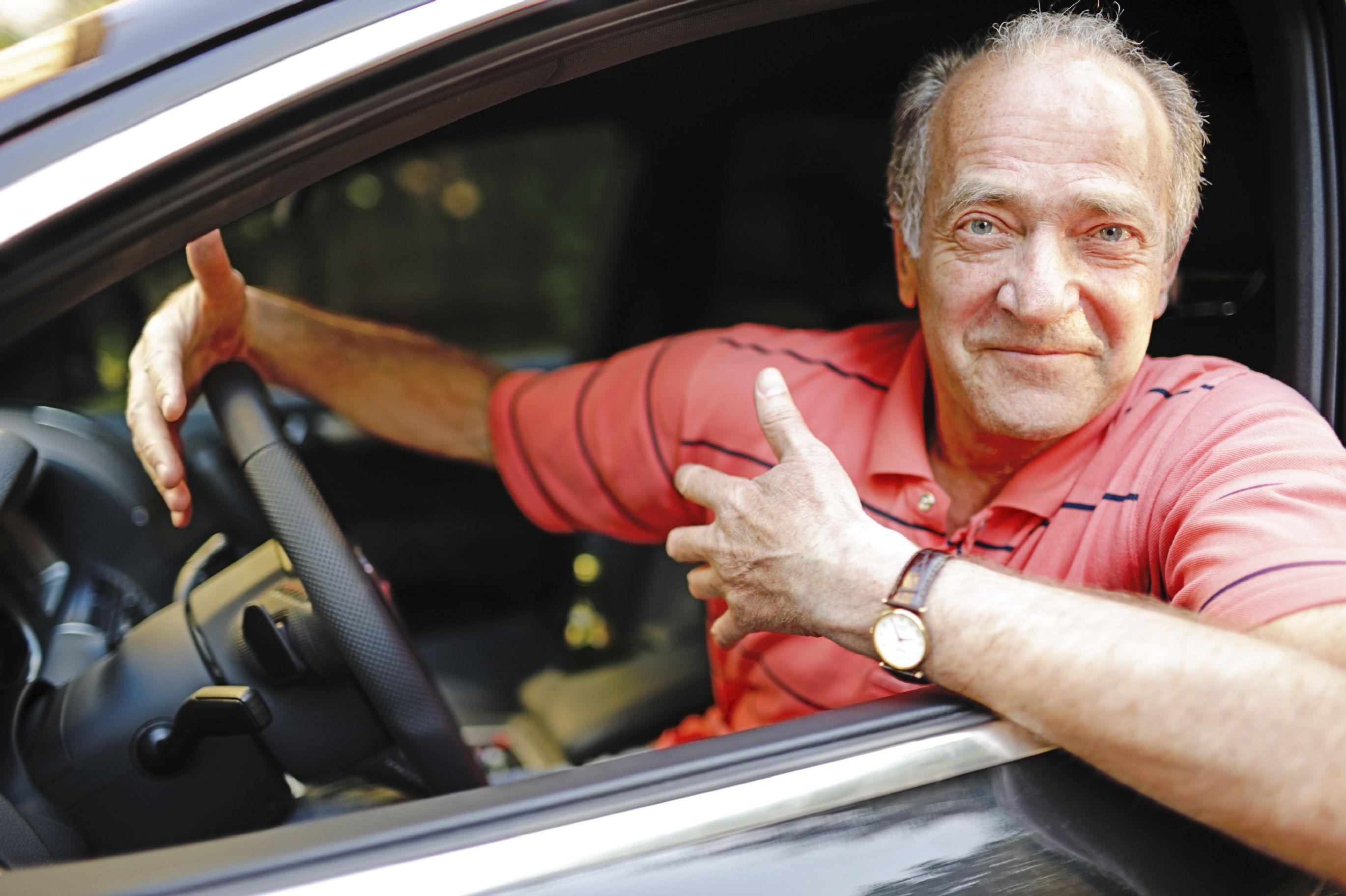 Man with receding hairline leaning out the window of a car smirking, wearing a watch with brown leather strap and a salmon coloured polo shirt with blue stripes.