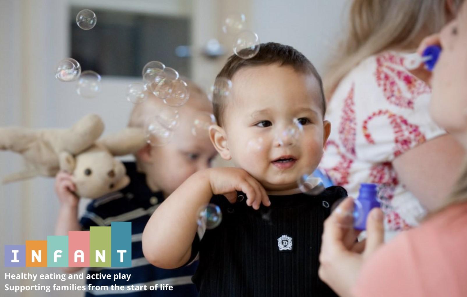 A smiling baby looks toward an adult who is blowing bubbles.