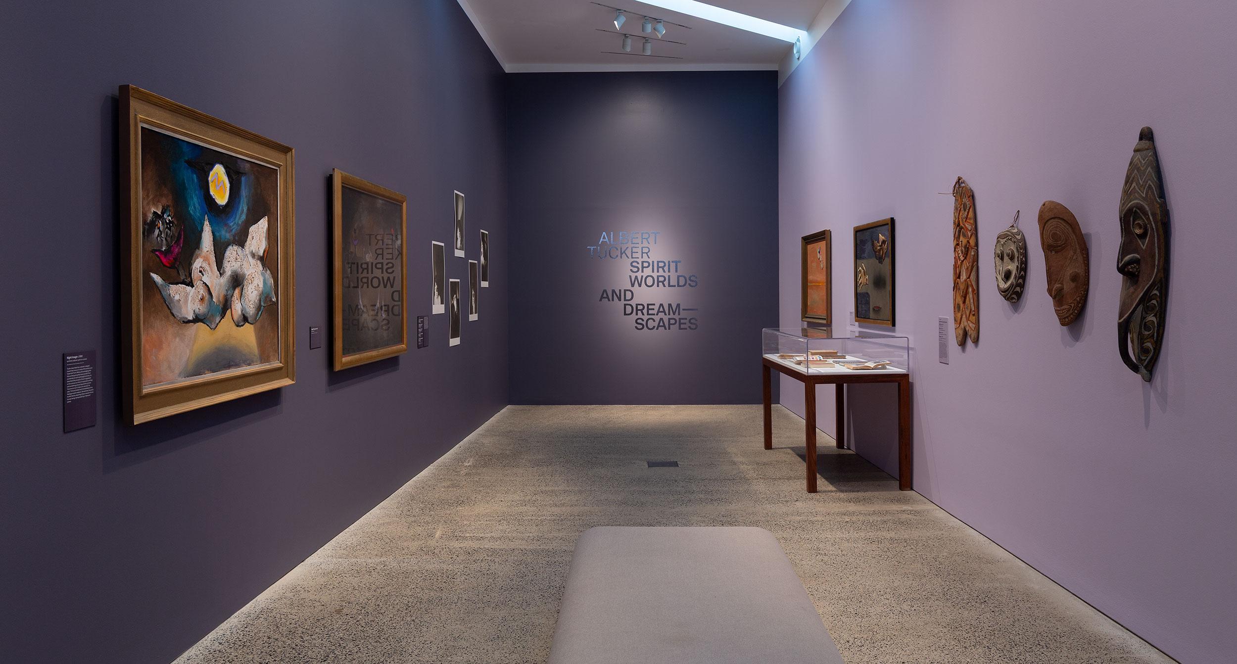 A small gallery room with purple walls. Paintings and sculptures hang on opposite walls and text is on the far wall.