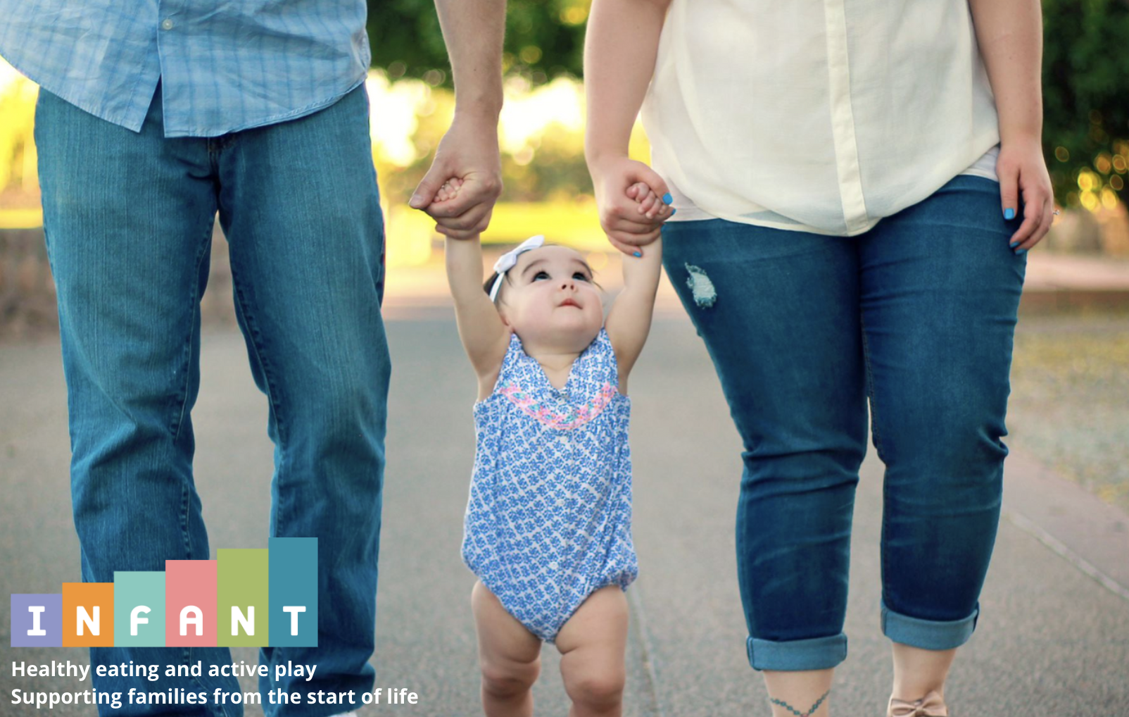 baby in between parents, holding their hands as they walk. Only the parents' legs are in view.