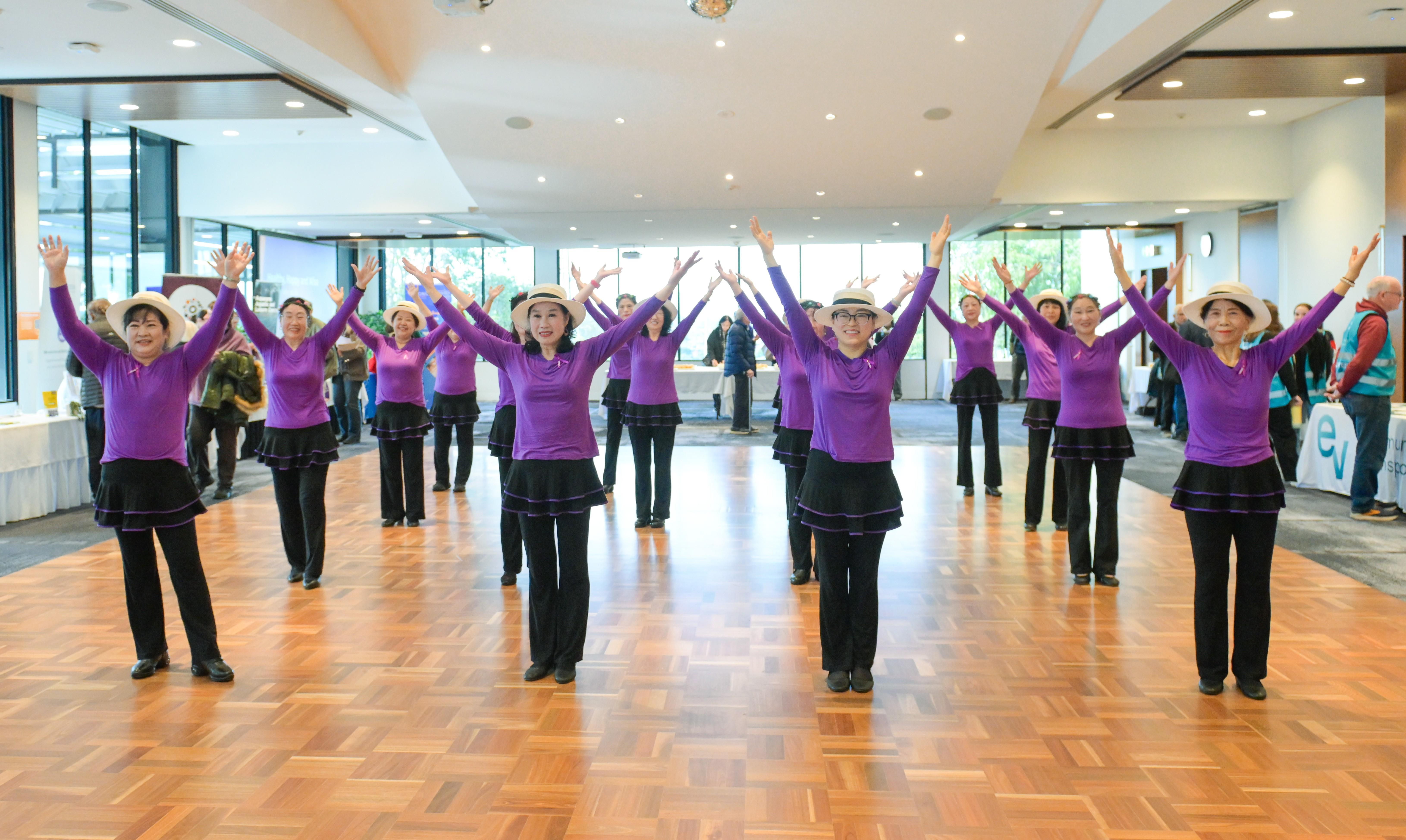 A group of dancers wearing purple tops, black dressings and pants and hats, from the Seniors Happy Life group, performing at the Manningham Function Centre.