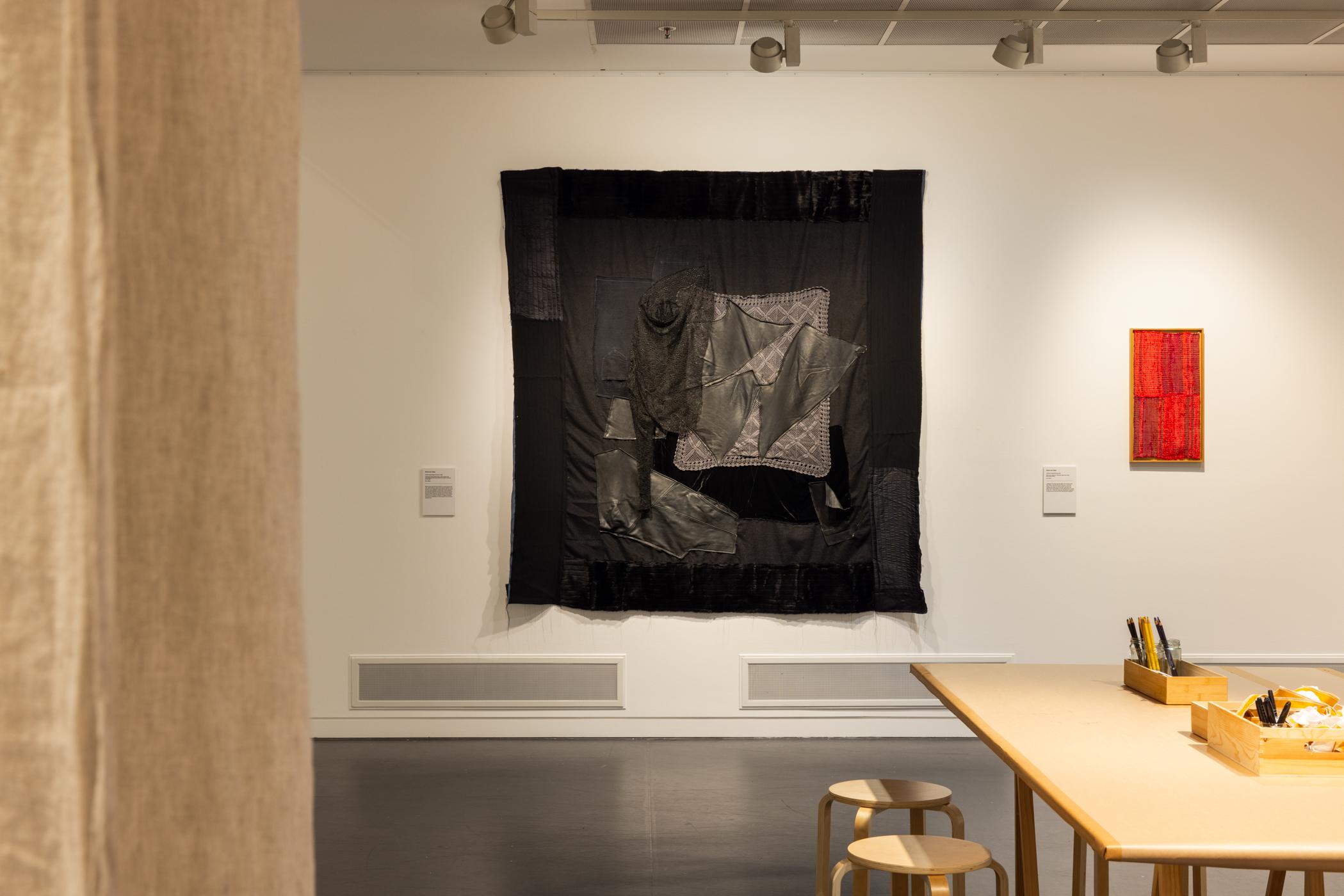 Gallery interior with a large black textile artwork on the wall, a smaller red piece to the right, and a wooden table with stools and art supplies in the foreground.