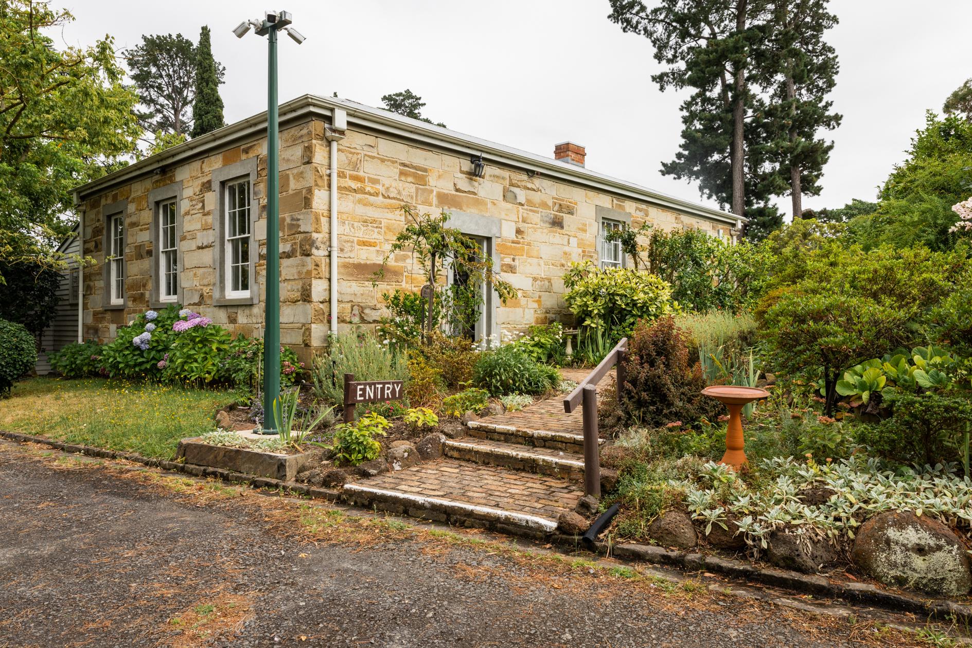 A restored sandstone building in a cottage garden setting. A small sign with the word 'entry' sits at the base of a path leading to the side door of the building.