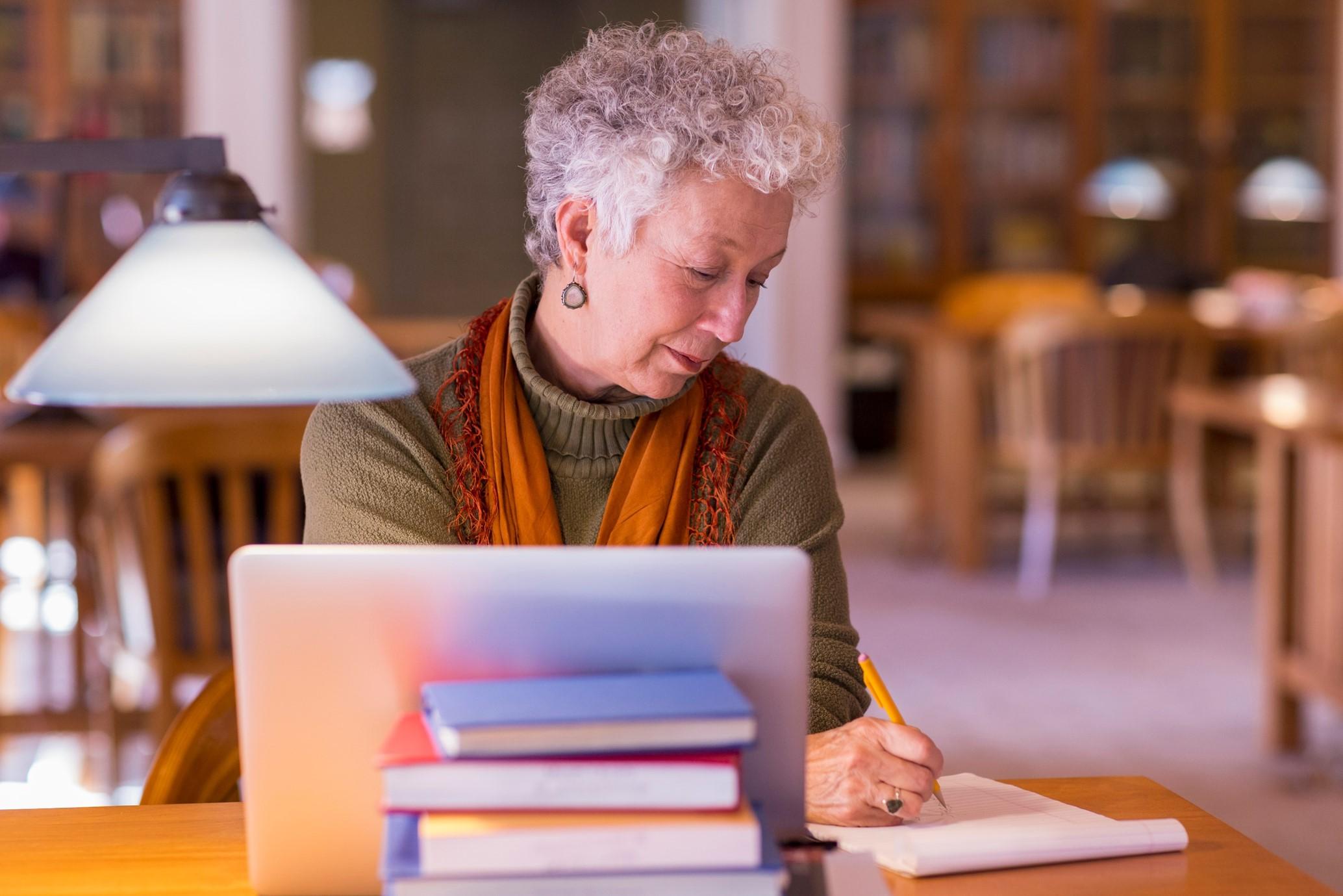 Older woman writing with pencil in front of a laptop and books