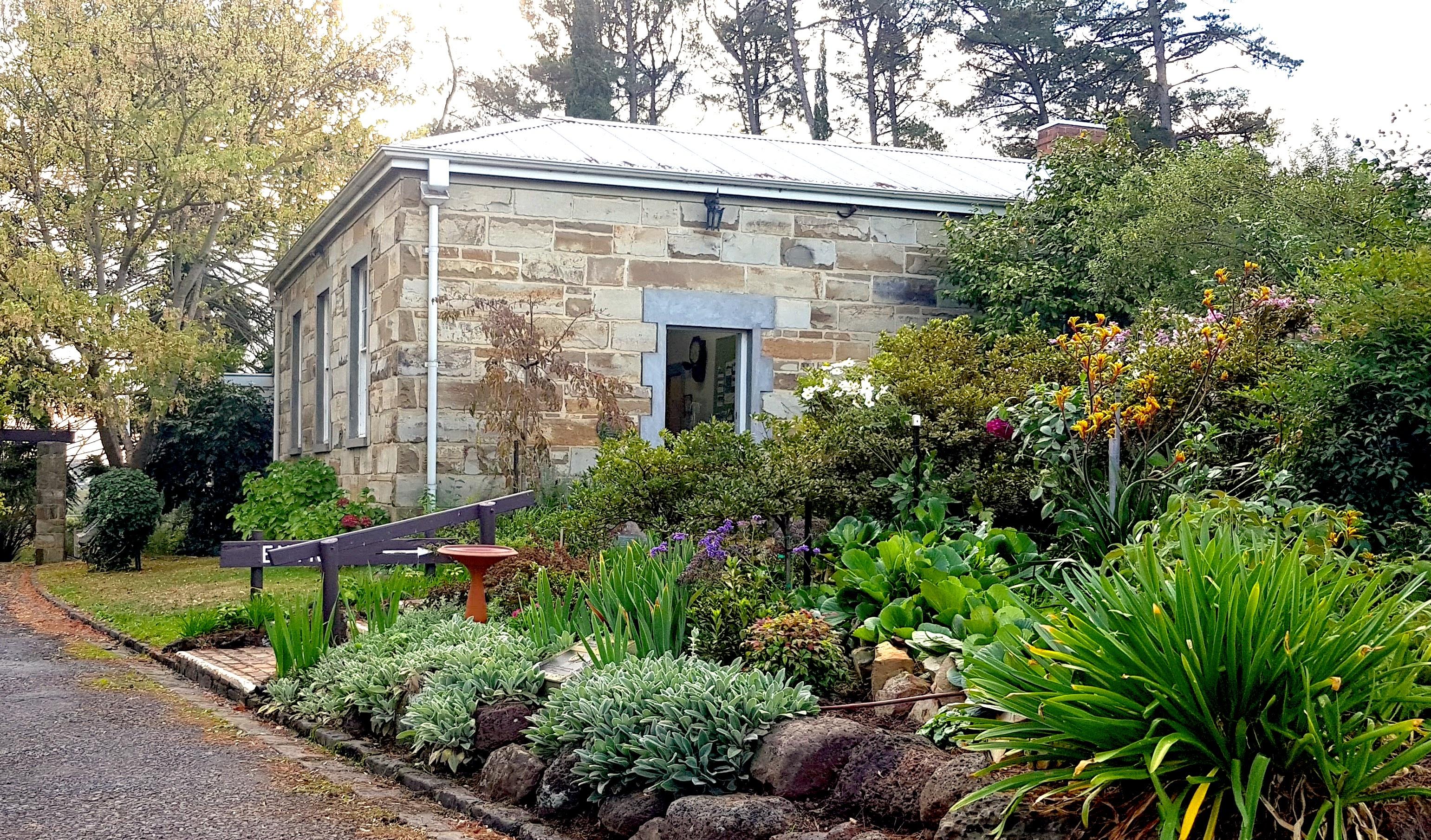 A historic stone cottage with a metal roof surrounded by lush greenery, colorful flowers, and shrubs along a stone-edged garden path, set against tall trees in the background.