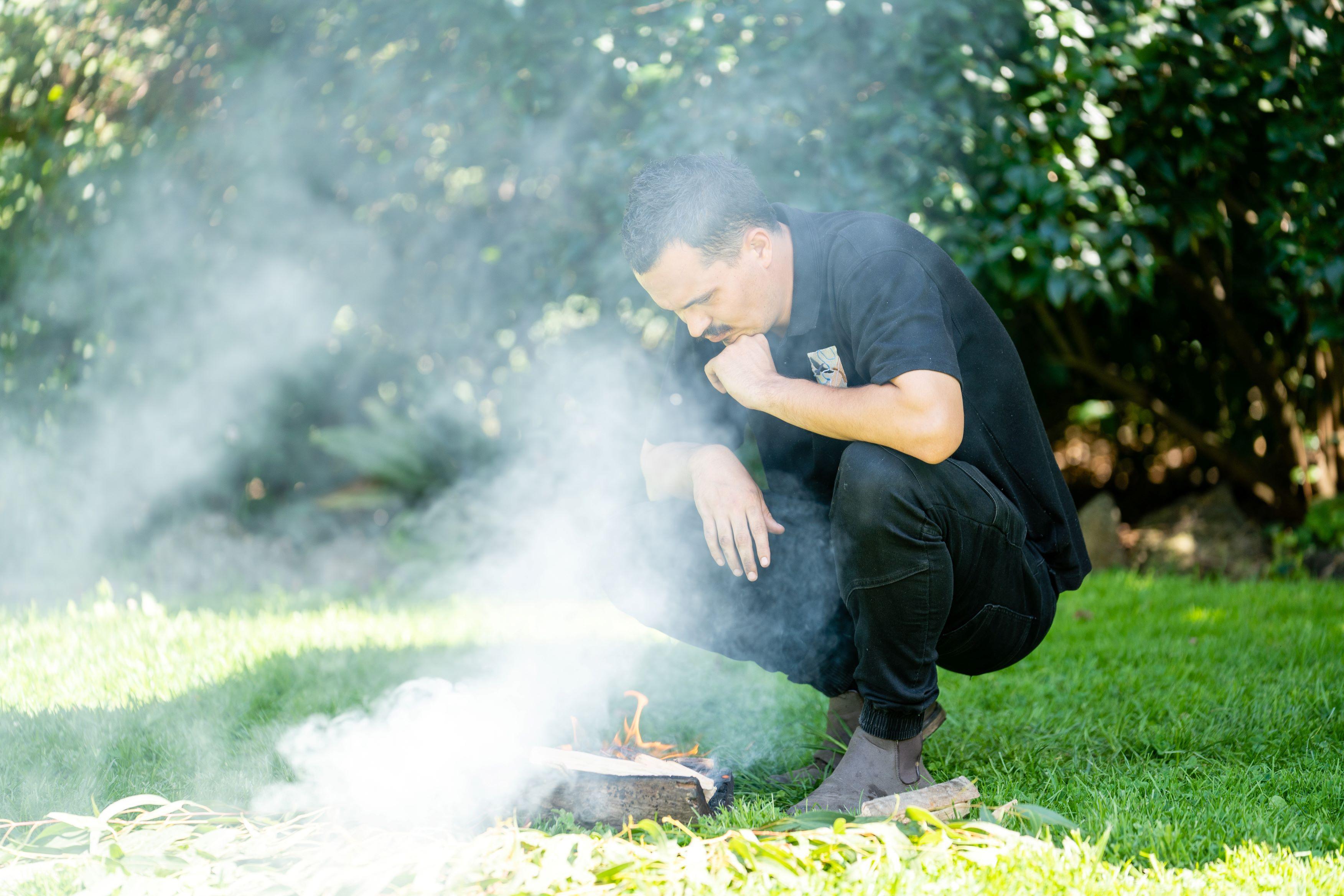 A young First Nations man dressed in black squats beside a small fire, smoke surrounds him and a lush green garden can be seen in the background.