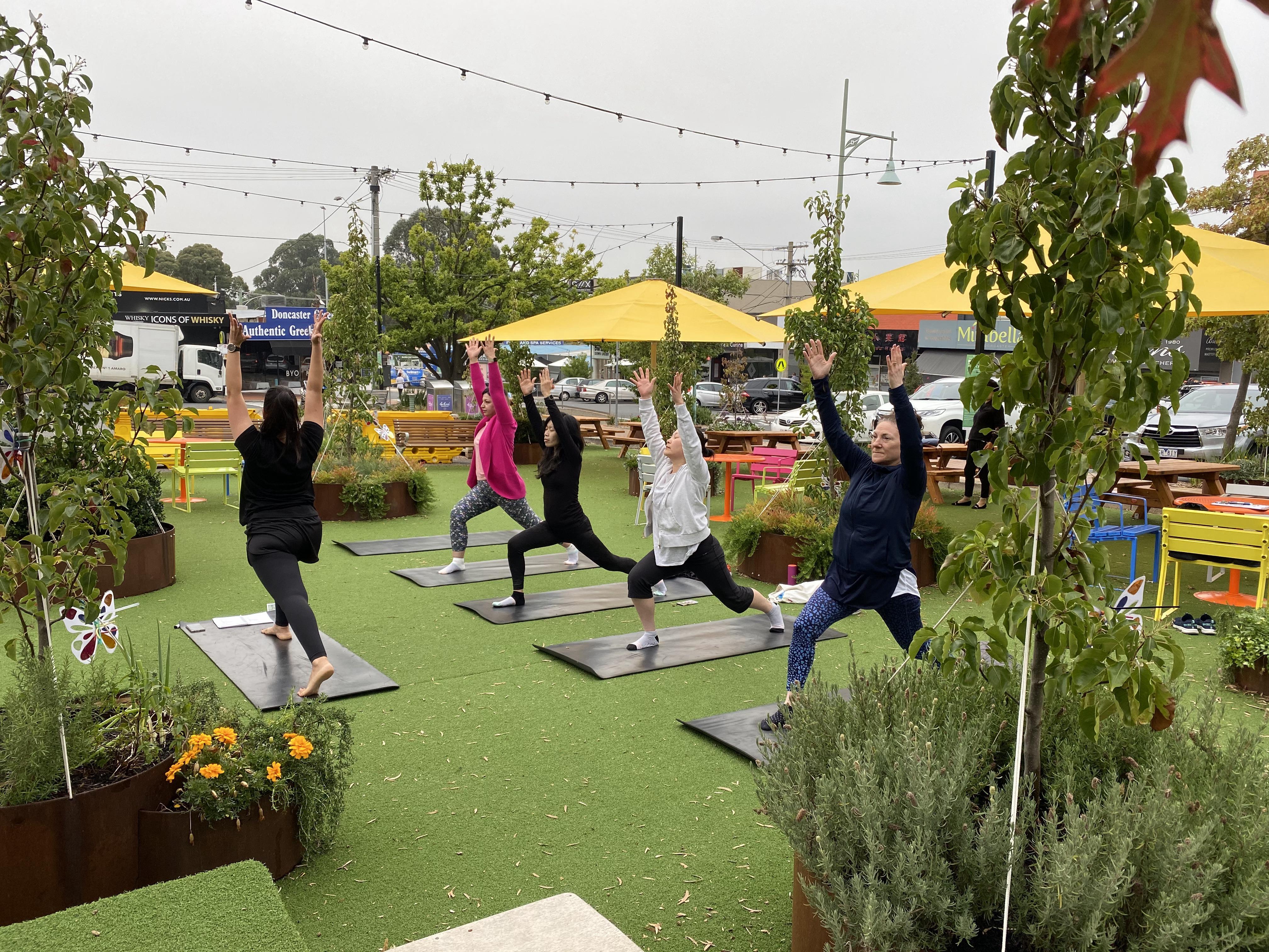 Outdoor yoga class with five people in warrior pose on black mats, surrounded by greenery and yellow umbrellas.