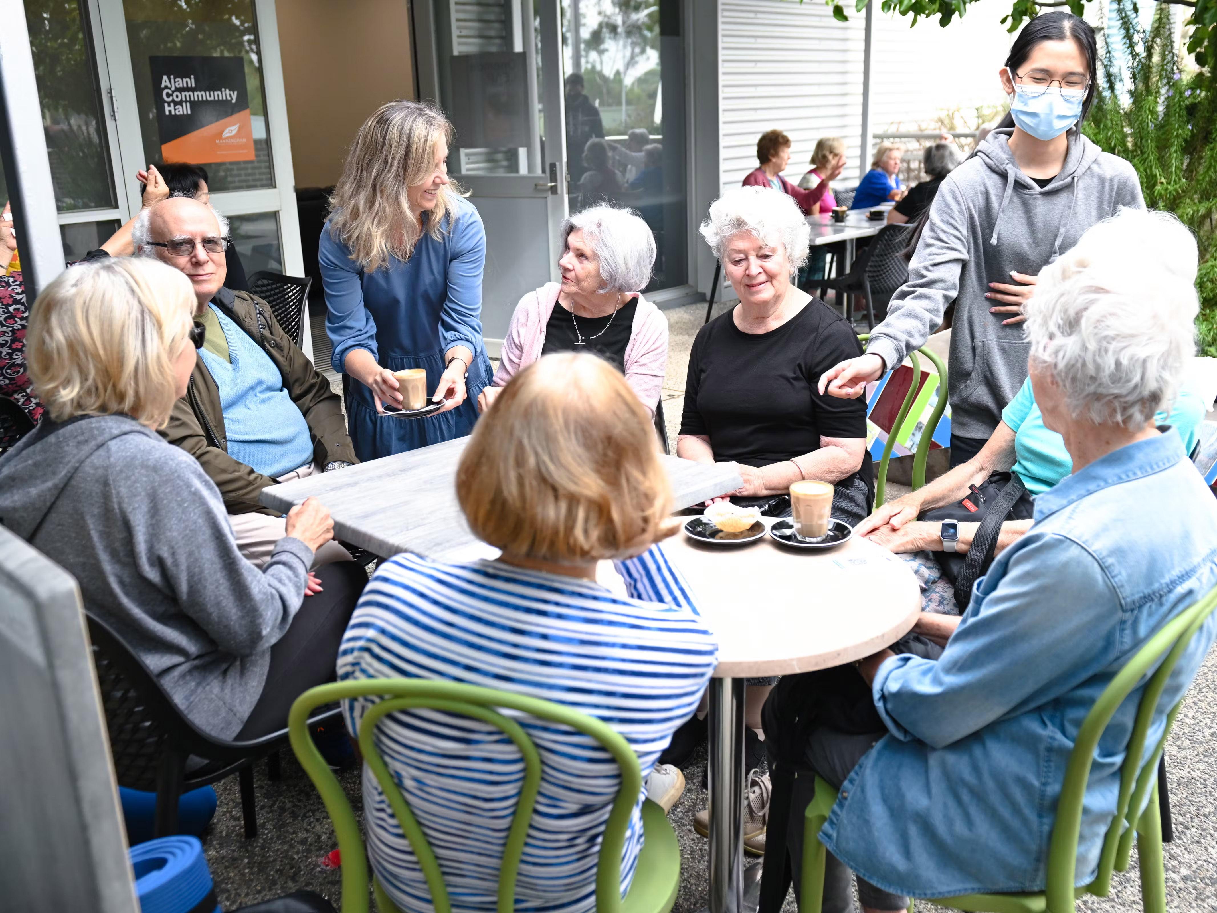 A group of older people sit around a table, they are being served coffee by two younger women. A sign with the words Ajani Community Hall can be seen on a window behind them.