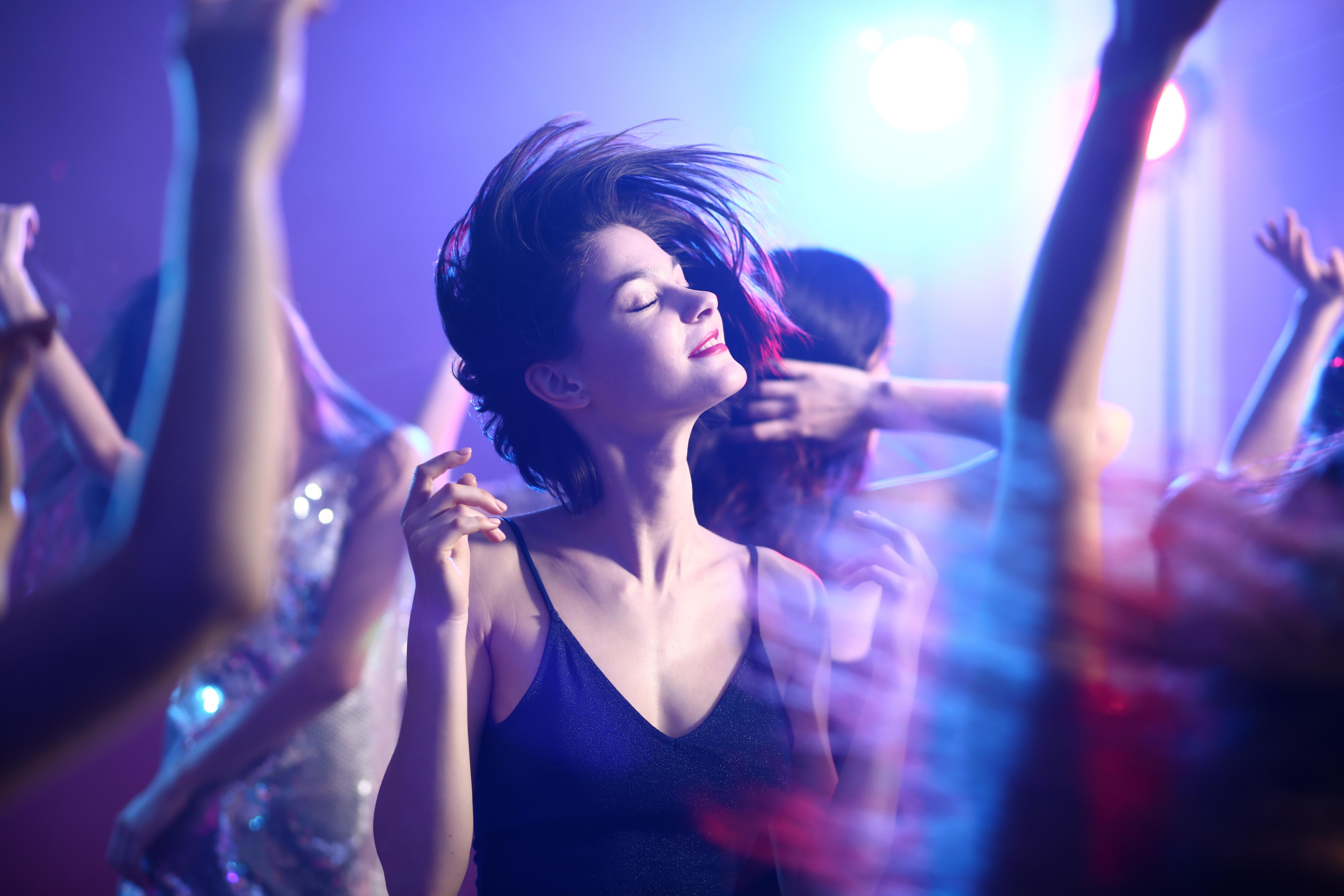Woman in a black dress with her eyes closed dances in a darkened room full of people
