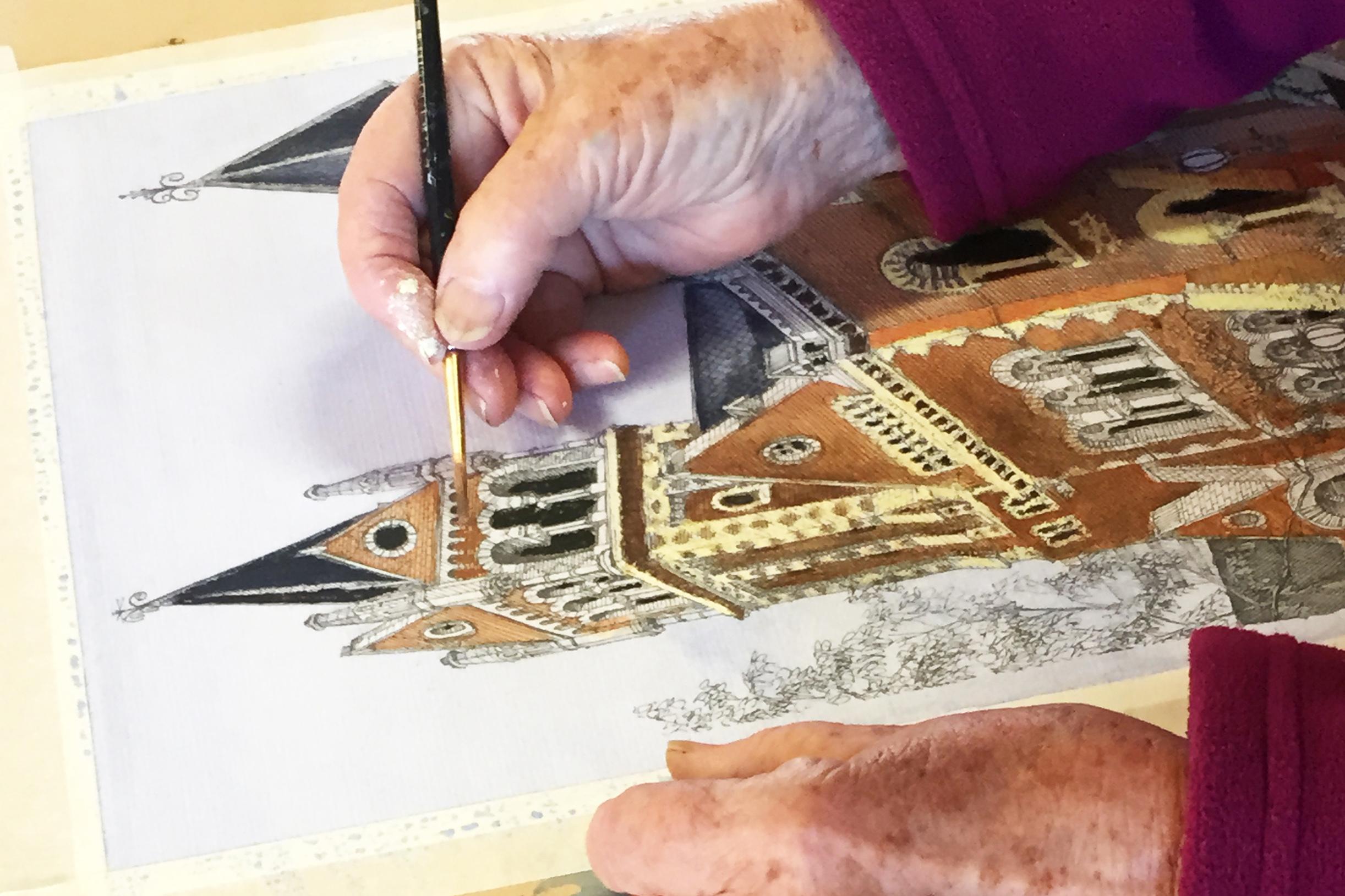 The hands of an older person, paint brush in one hand, resting over a painting of a brown brick building with an elaborate steeple.