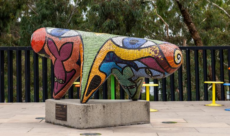 Sculpture of an abstract three-legged creature covered in small colourful mosiac tiles mounted on a concrete block, gum trees in the background.