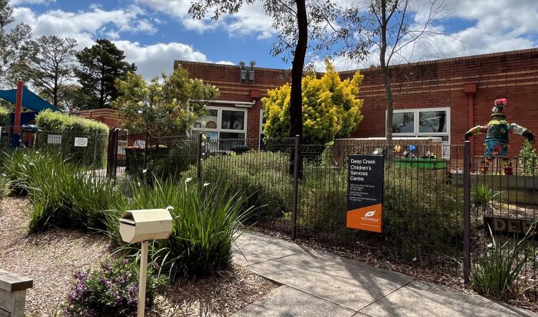 A low red brick building surrounded by a shrubby garden and a wire front fence.