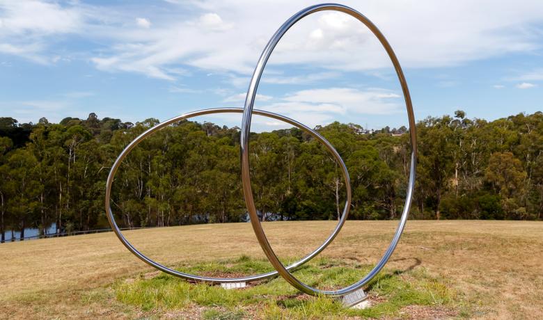 Two large polished stainless steel circles sit on a hill overlooking an open grassy expanse with gum trees and a lake in the background.