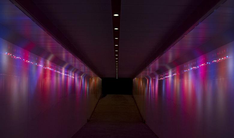 Red, purple and blue waves of light run vertically along the length of a dark underpass, a strip of small white circular lights runs along the ceiling