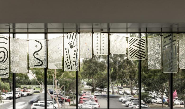 A series of 12 glass panel of different sizes, all with unique markings and textures, joined together in a wave-like pattern. A parking lot with cars and gum trees can be seen through a window behind the panels.