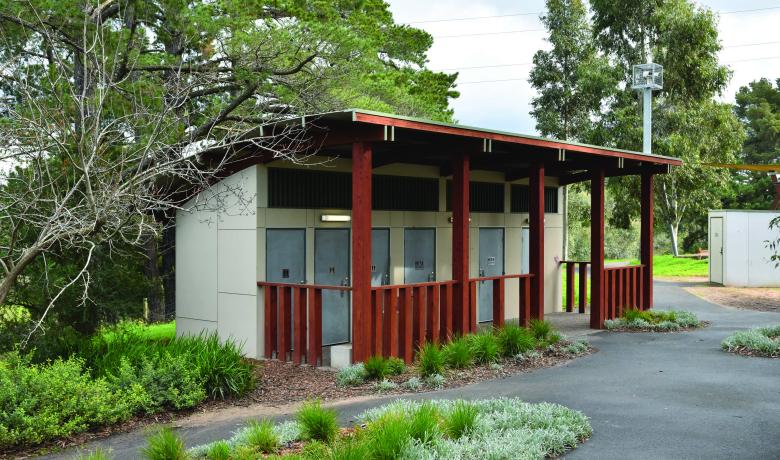 A block of six public toilets with a small wooden porch in front, set in a bushy park with paths winding between trees.