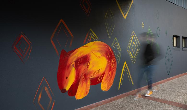 Mural of an orange and red wombat on a grey wall, a blurred figure walks in front of the wall.