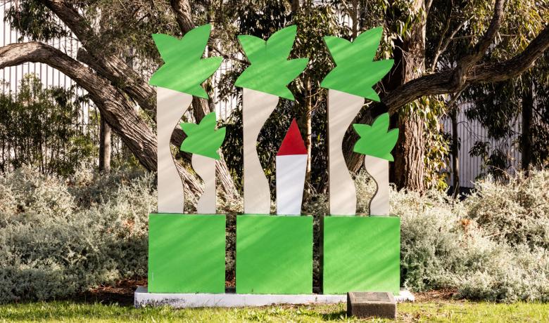 A two dimensional concrete sculpture sits in a garden of native plants with a steel fence in the background. The sculpture is in the shape of five green and brown trees of differing heights and a small red and white rocket in the centre,