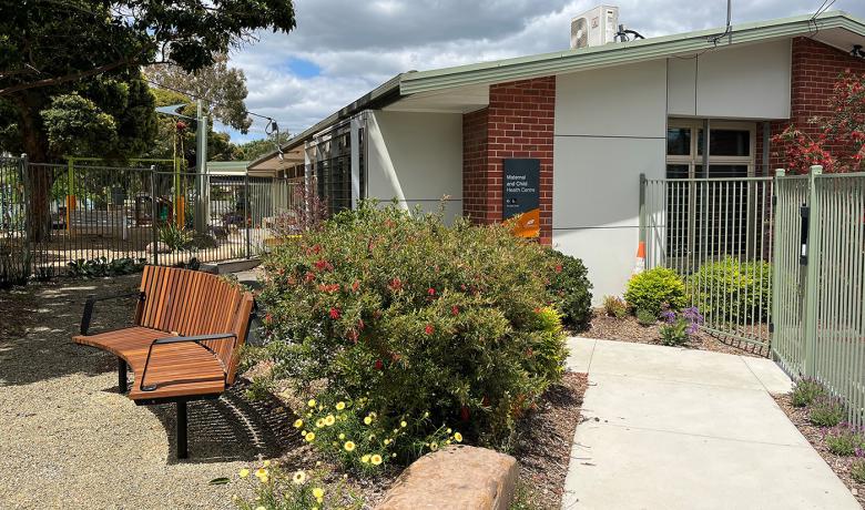 A red and white brick building surrounded by a garden with a bench set and concrete paths.