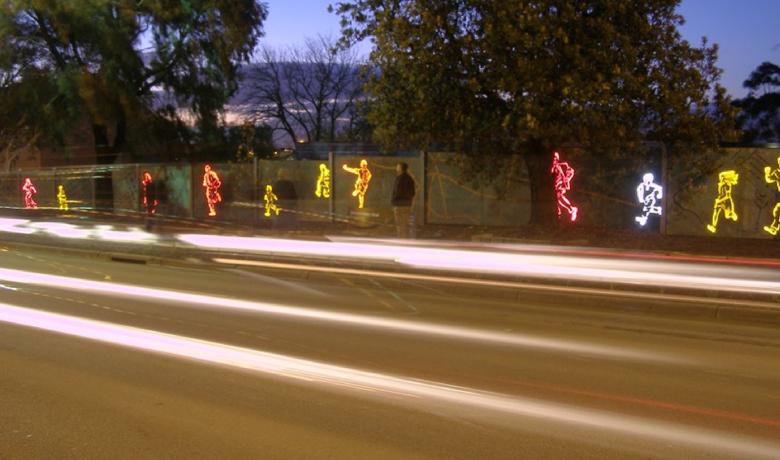 Illuminated human figures in pink, yellow and white appear on a wall alongside a road at dusk