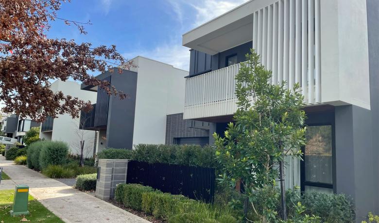 A row of modern, two-storey town houses on a residential street. Small trees and shrubs line the footpath.