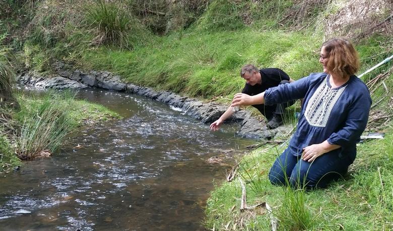Two people kneel by the edge of a river flowing through bushland