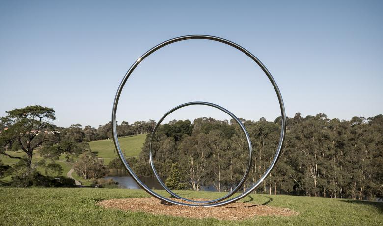 Two large polished stainless steel circles sit on a hill overlooking an open grassy expanse with gum trees and a lake in the background.