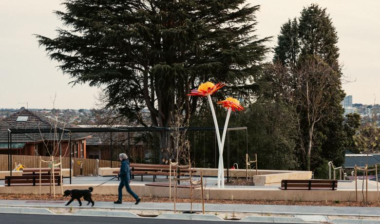 A woman walks her dog in front of a contemporary urban park with large trees in the background. At the centre of the park is a large sculpture which takes the form of two orange flowers on long stems.