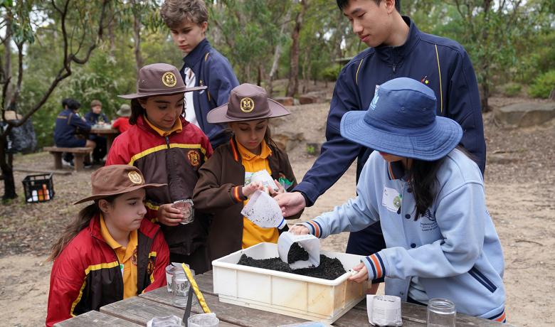 A group of kids of mixed ages and genders stand at a wooden table in a bushy environment. They are scooping dirt into plastic containers.