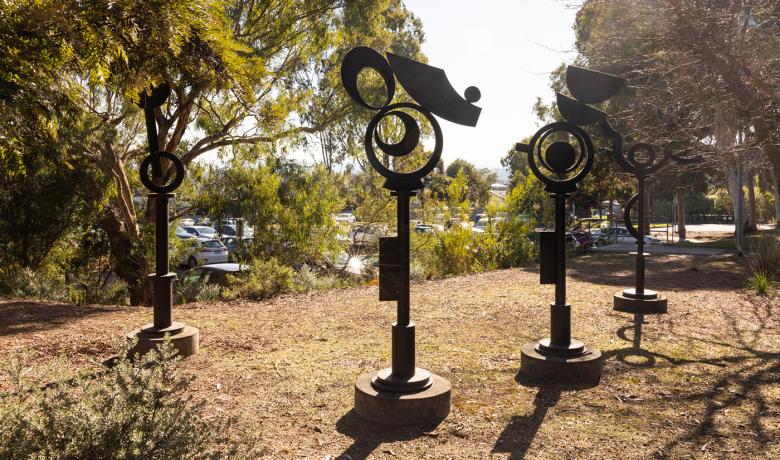 Four tall totemic steel sculptures sit in a clearing amongst bushy native trees, a carpark is in the background.