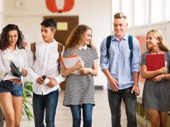 Five young adults carrying books, walking down a wide hallway, smiling and looking at each other