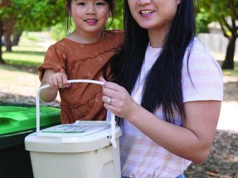 A young woman is holding a child on her hip, together they are holding a light brown FOGO caddy