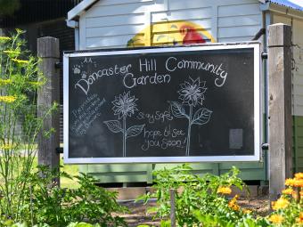 A chalkboard sign sits amongst a veggie garden with a colourful mural in the background. The sign says 'Doncaster Hill Community Garden. Stay safe. Hope to see you soon"