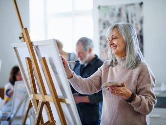 A woman with long grey hair standing at an easel painting in a class with other participants in the background.