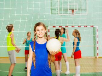 Girl holding a volleyball standing in front of a volleyball net in a stadium.