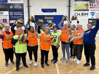 Group of 11 women standing on a basketball court each holding a basketball in the air and wearing high-vis basketball vests with their coach on the right