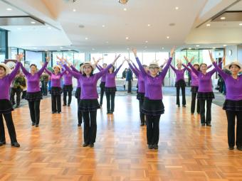 A group of dancers wearing purple tops, black dressings and pants and hats, from the Seniors Happy Life group, performing at the Manningham Function Centre.