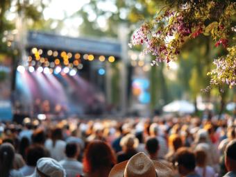 Photograph showing a large group of people from behind watching a performance on a stage in the distance. It is daytime but there are colourful lights on the stage and beaming out across the audience.