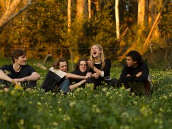 Members of the band BOTH sitting in a field of grass and yellow flowers with bushland in the background.