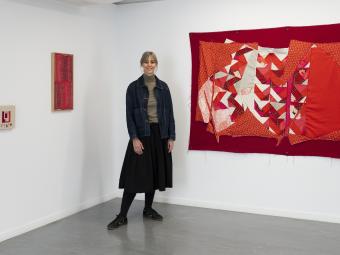 Artist Shannon Slee, wearing a black skirt and denim jacket, stands in the corner of a room next to a large red tapestry which is hanging on the while walls on the right, and a small framed red tapestry on the left.